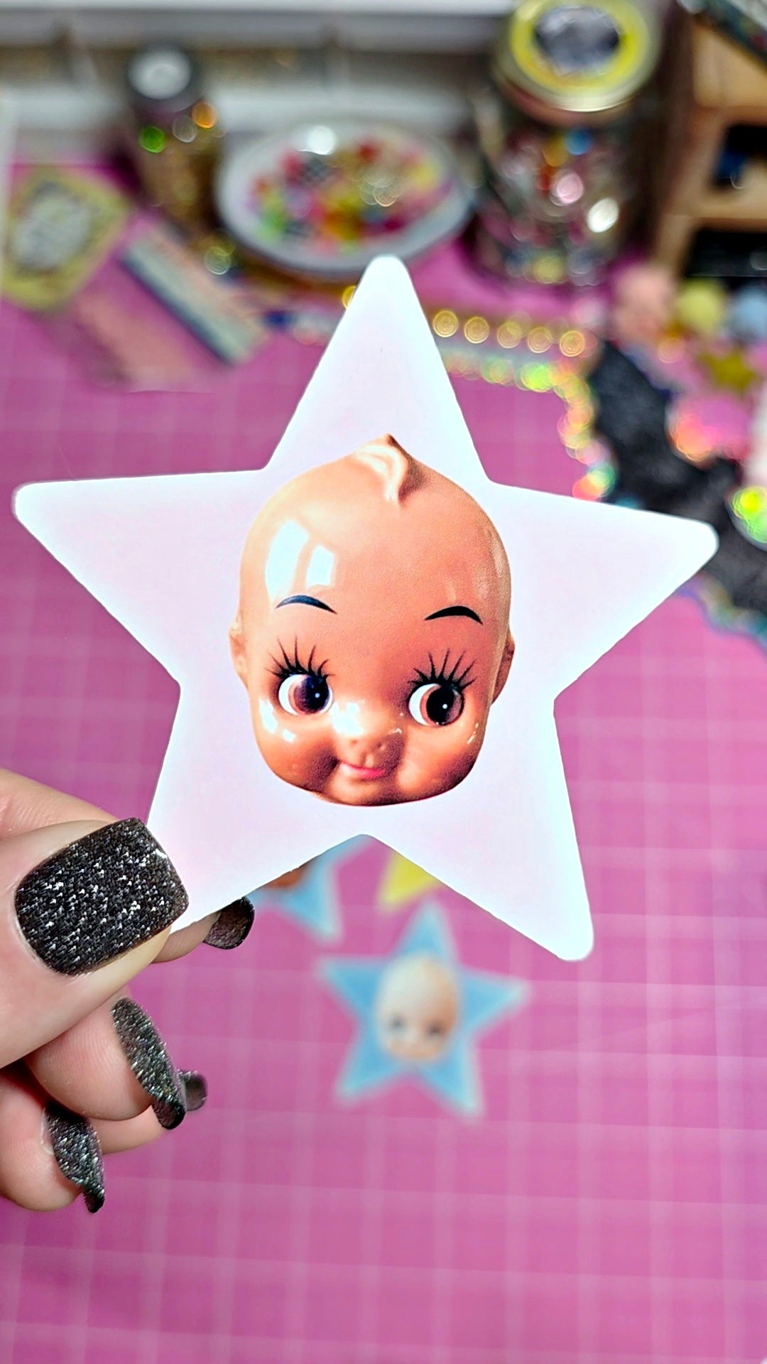 A hand with glittery black fingernails holds a white star-shaped sticker or card featuring a vintage-style doll face with large eyes and long eyelashes. The background is a pink cutting mat with other colorful star-shaped items and craft supplies blurred behind it.