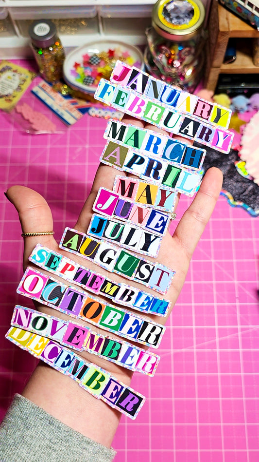 A hand holds a vertical stack of colorful paper strips printed with the months January through December, set against a pink cutting mat and craft supplies in the background.