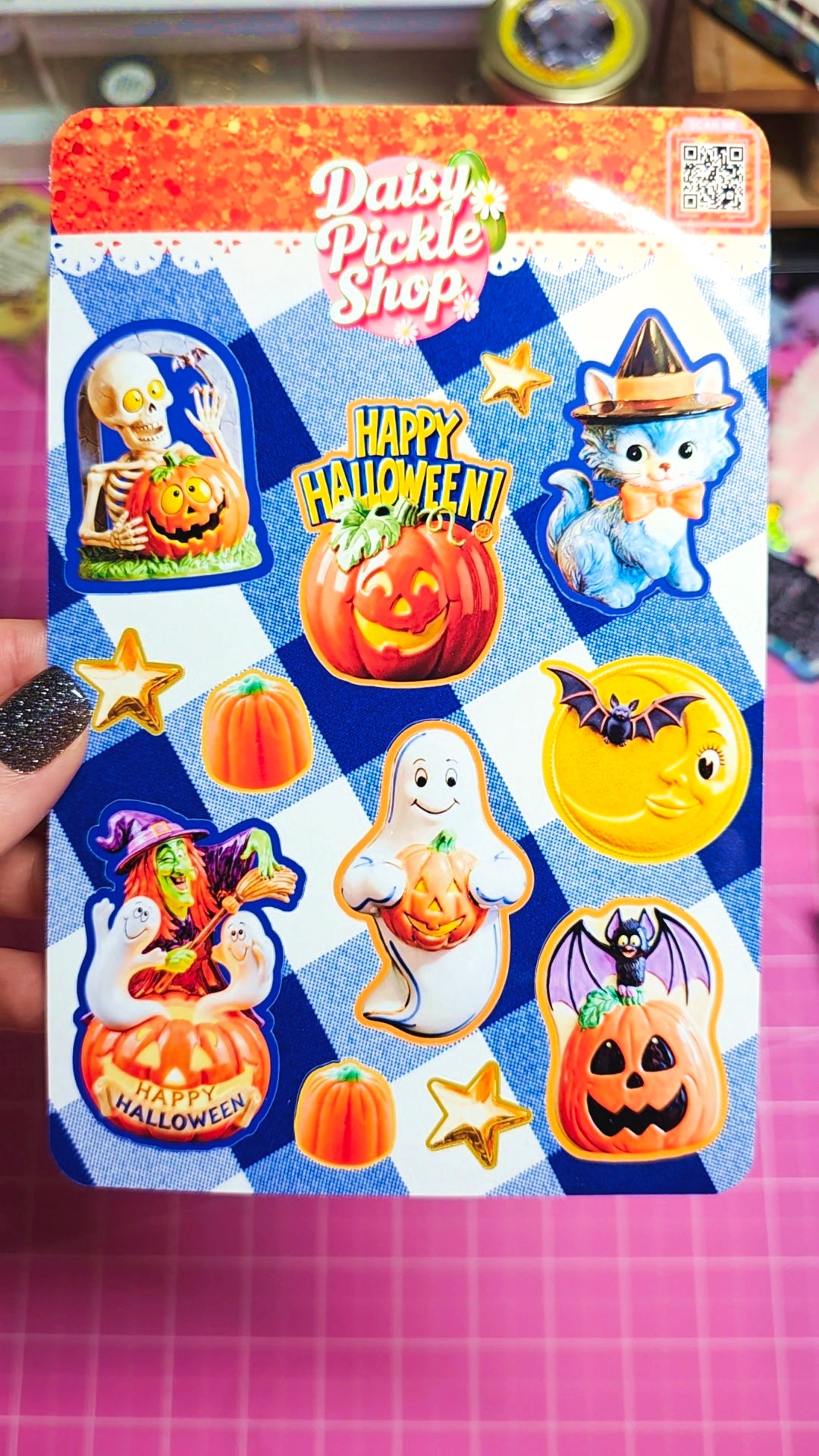 A hand with glittery black nail polish holds a sticker sheet from Daisy Pickle Shop featuring various Halloween-themed stickers, including pumpkins, ghosts, a cat in a witch hat, and a moon, on a blue and white checkered background.