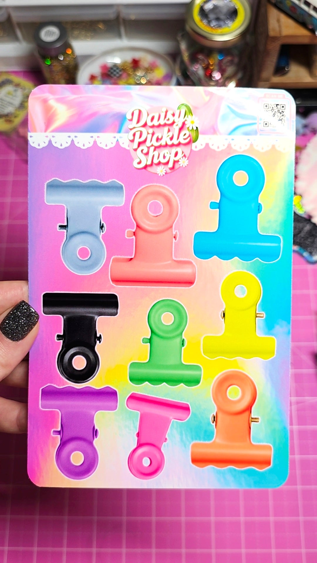 A hand holds a package of eight colorful plastic binder clips in various shapes and sizes. The packaging features a pastel rainbow gradient background with the brand name 'Daisy Pickle Shop' printed at the top.