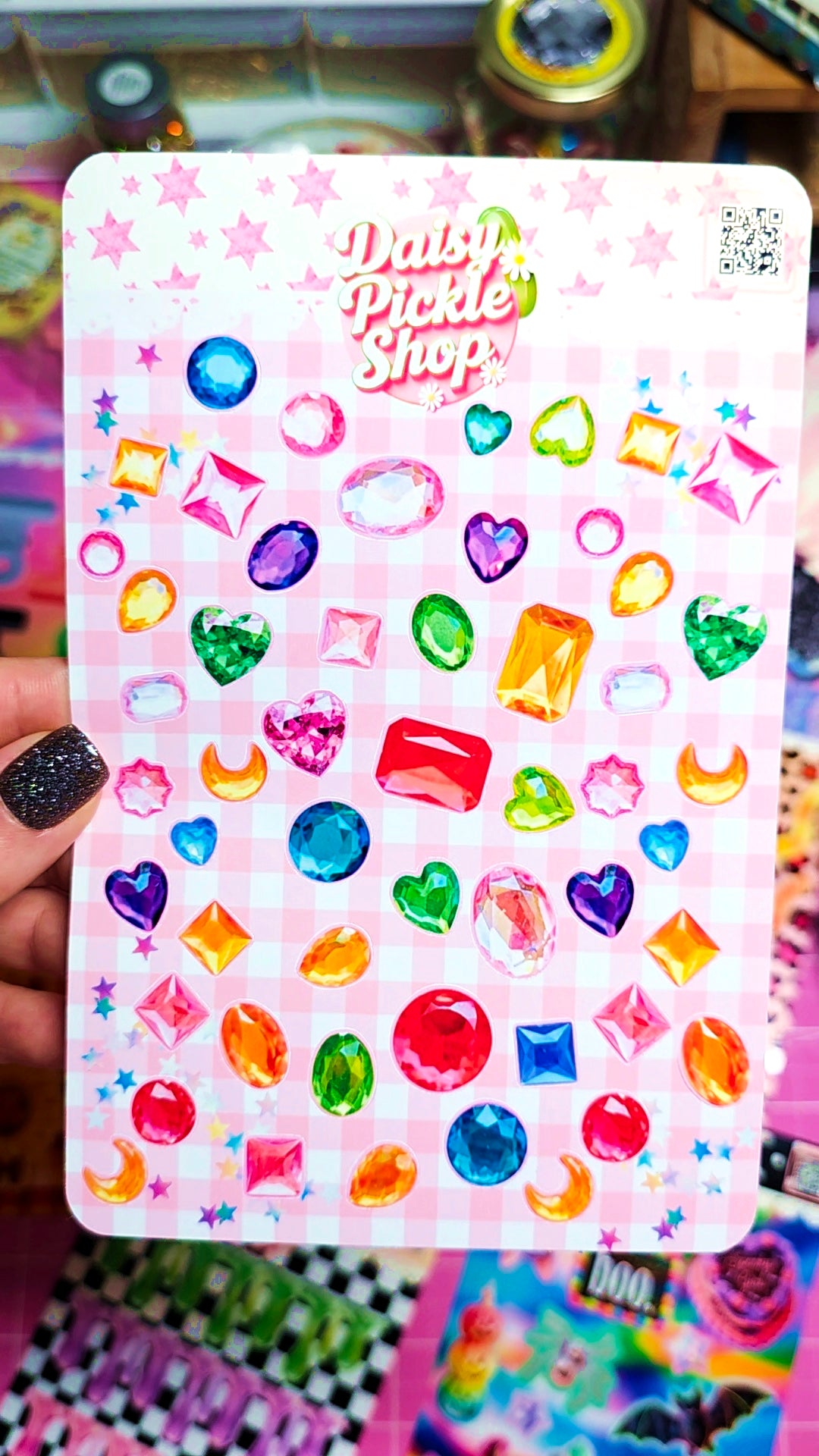 A hand with black glitter nail polish holds a sticker sheet from Daisy Pickle Shop. The sheet features a pink gingham background decorated with colorful, shiny gem shapes like hearts, diamonds, crescents, and stars.