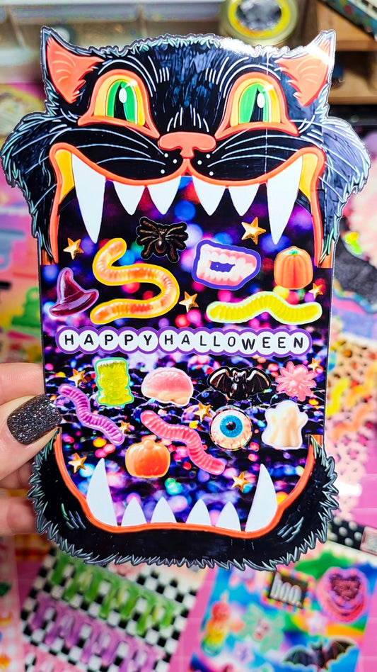 A hand with glittery black nail polish holds a die-cut Halloween sticker featuring a black cat face with glowing green eyes and an open mouth full of candy and spooky illustrations, with 'HAPPY HALLOWEEN' text across the center.
