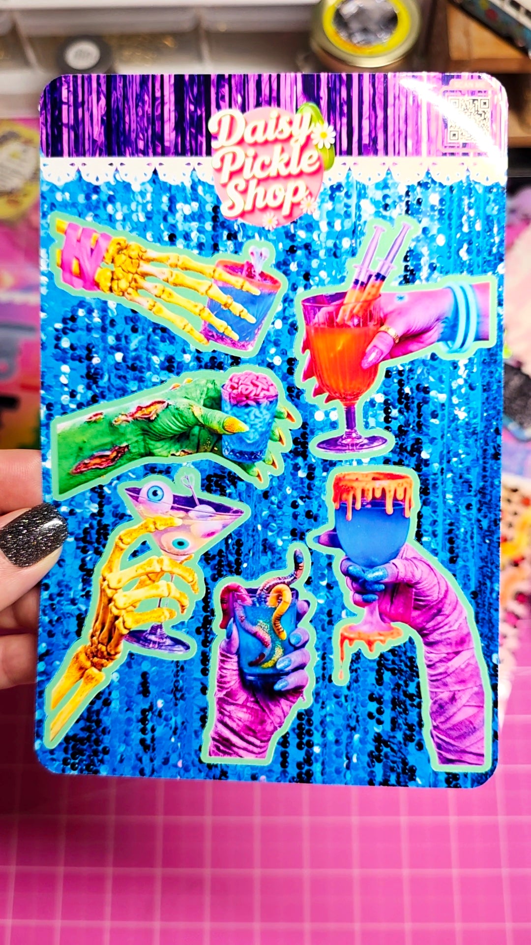 A hand holds a Dashes Pickle Shop sticker sheet featuring colorful neon designs of skeletal hands holding drinks and food, set against a blue sequined background.