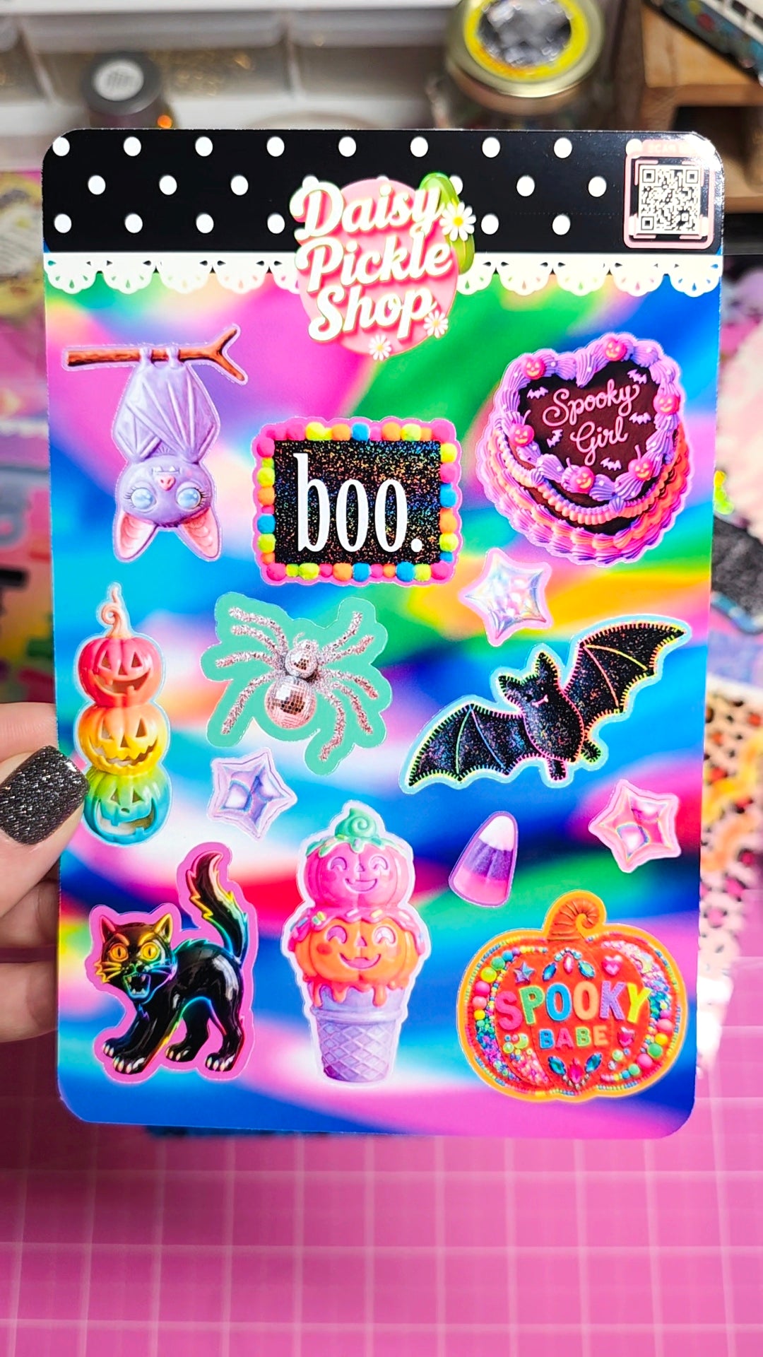 A hand holds a packaged set of colorful Halloween-themed stickers from Daisy Pickle Shop. The sticker sheet features spooky designs including bats, a black cat, jack-o'-lanterns, a spider, a ghost, and candy decorations on a bright rainbow background.