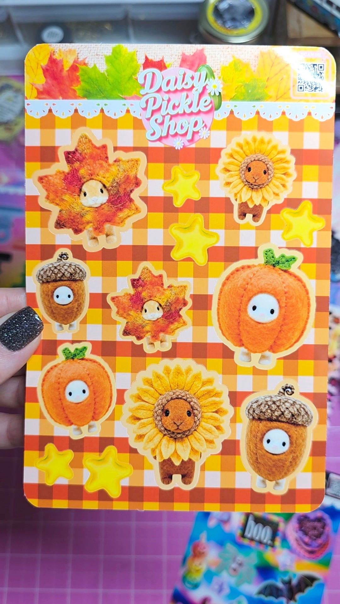 A hand with black glitter nail polish holds a sticker sheet from Daisy Pickle Shop. The sheet features cute autumn-themed characters including sunflowers, pumpkins, acorns, and maple leaves on a yellow and orange gingham background, with yellow stars.