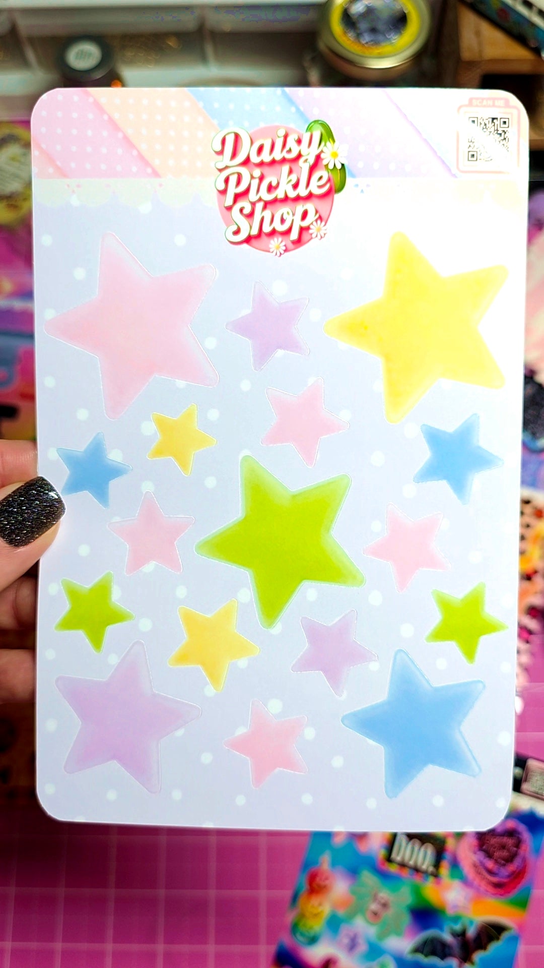 A hand with black glitter nail polish holds a sticker sheet from Daisy Pickle Shop. The sheet features a white background decorated with pastel pink, yellow, blue, and green star stickers.