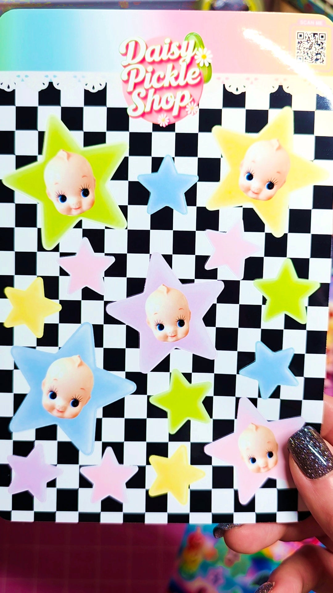 A hand holds a sticker sheet featuring retro-style baby face stickers inside pastel-colored star shapes on a black-and-white checkered background. The sheet has a 'Daisy Pickle Shop' logo at the top and glittery dark nail polish is visible on the holding finger.
