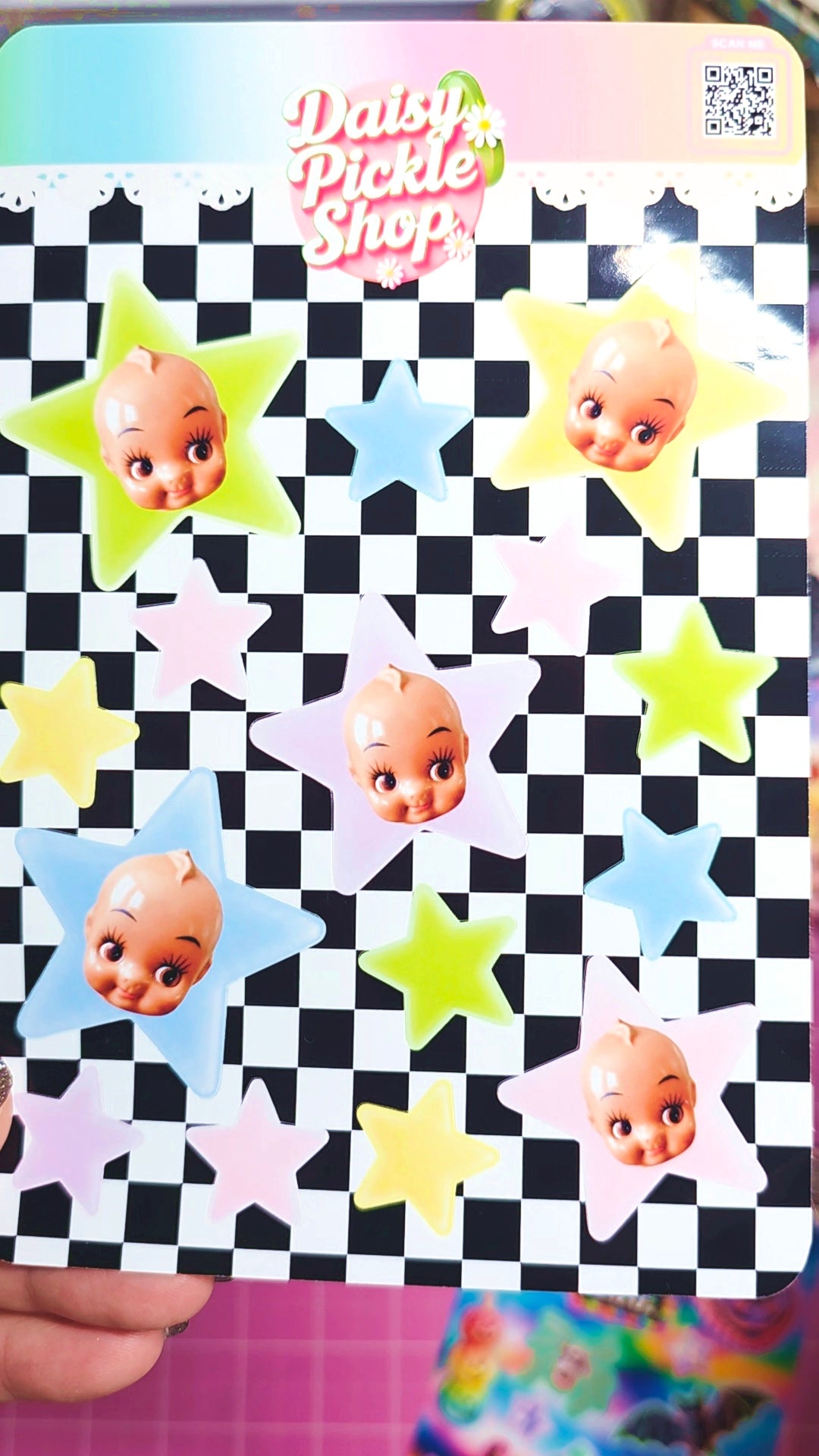 A hand holds a printed sticker sheet featuring cartoon baby faces inside pastel-colored stars over a black-and-white checkerboard background. The sheet has a pink header that reads 'Daisy Pickle Shop' and a QR code in the top right.