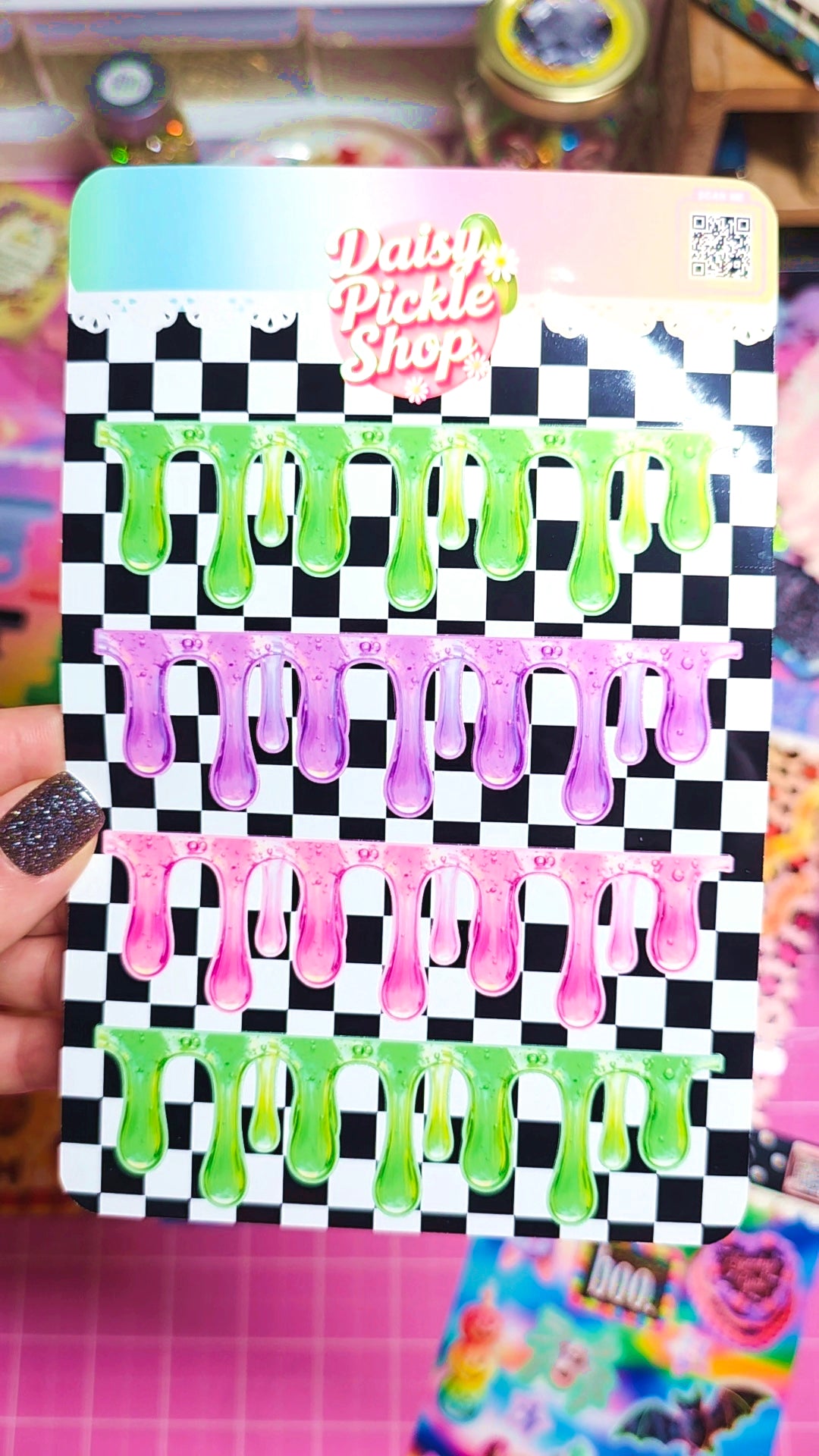 A hand holds a card of Daisi Pickle Shop nail wraps featuring four rows of dripping slime designs in green, pink, and purple on a black and white checkered background.