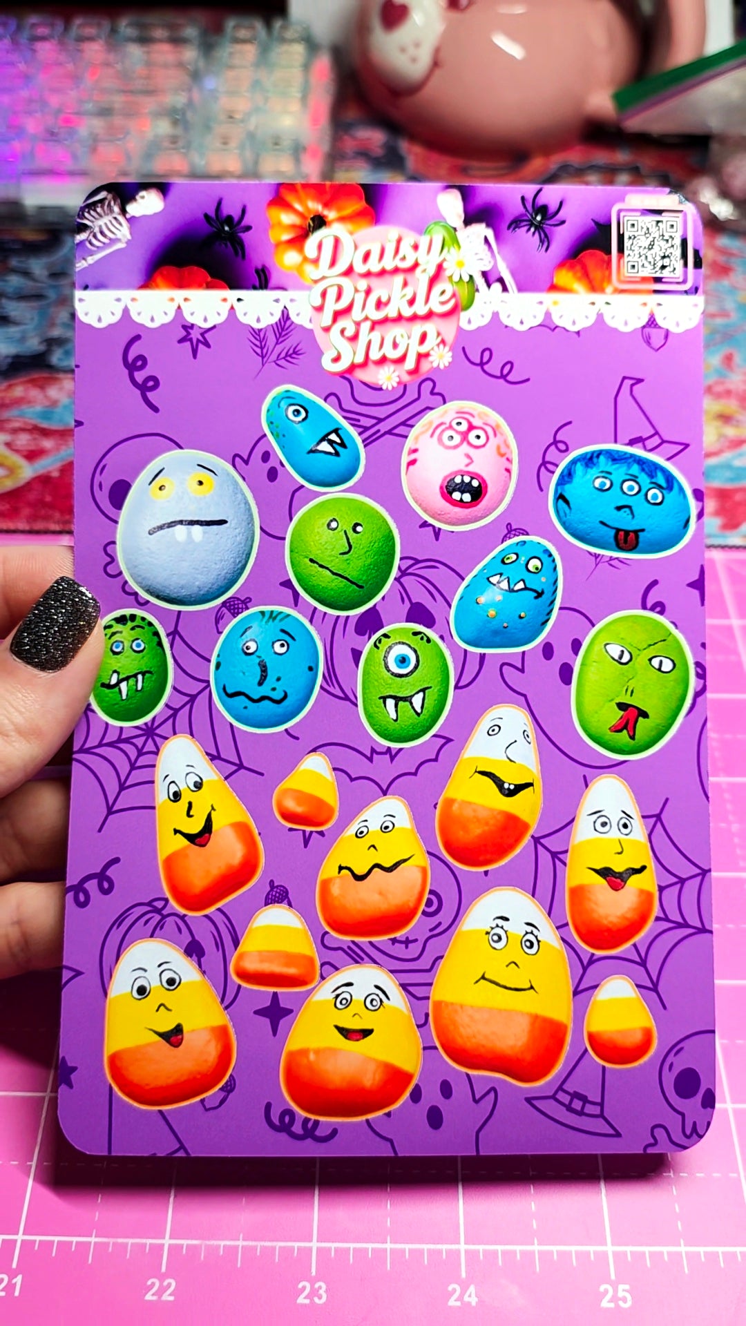 A hand with black glitter nail polish holds a purple card featuring colorful candy corn and round monster faces, branded 'Daisy Pickle Shop,' set against a pink cutting mat.