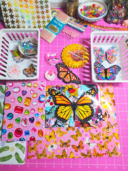 A flat lay of colorful craft supplies and decorative items arranged on a bright pink cutting mat, including butterfly-themed sheets, jewel stickers, white baskets with embellishments, and small packaged accessories.