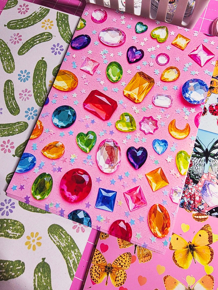 A close-up of pink craft or scrapbook paper decorated with colorful faux gem stickers, alongside other patterned papers featuring cacti and butterflies.