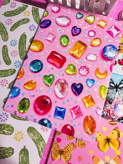 A close-up of pink craft or scrapbook paper decorated with colorful faux gem stickers, alongside other patterned papers featuring cacti and butterflies.