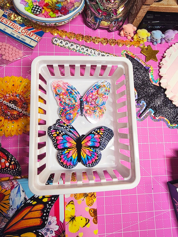 Three colorful butterfly-shaped resin charms displayed inside a white plastic laundry basket, set on a pink cutting mat surrounded by craft supplies.
