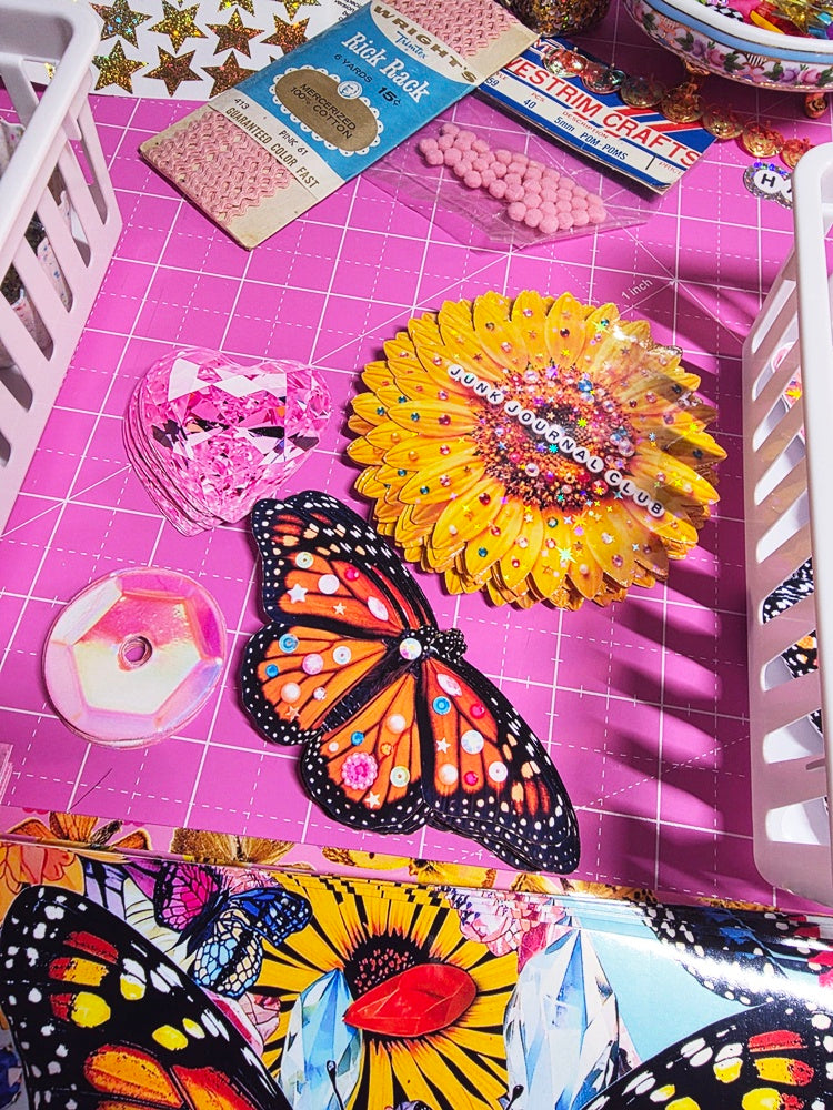 A collection of colorful craft supplies and decorative items arranged on a pink grid cutting mat inside a white basket. Visible items include a butterfly-shaped ornament, a yellow flower with beads, pink sequins, a pink heart-shaped embellishment, a spool of pink thread, and packaged beads and ribbon.