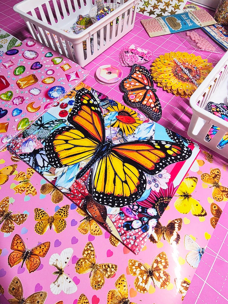 A colorful craft or stationery set featuring a large printed butterfly design on a pink sheet, surrounded by smaller butterflies, rhinestone stickers, floral paper cutouts, and white plastic baskets on a pink cutting mat.