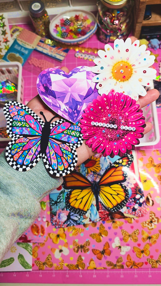 A hand holding several colorful butterfly and flower-themed stickers or craft embellishments over a pink patterned surface with more decorative items in the background.