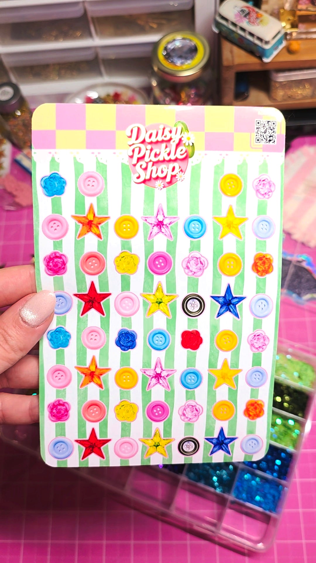 A hand holds a card of colorful craft buttons and star-shaped embellishments under a 'Daisy Pickle Shop' logo. The background shows a pink cutting mat and storage bins.