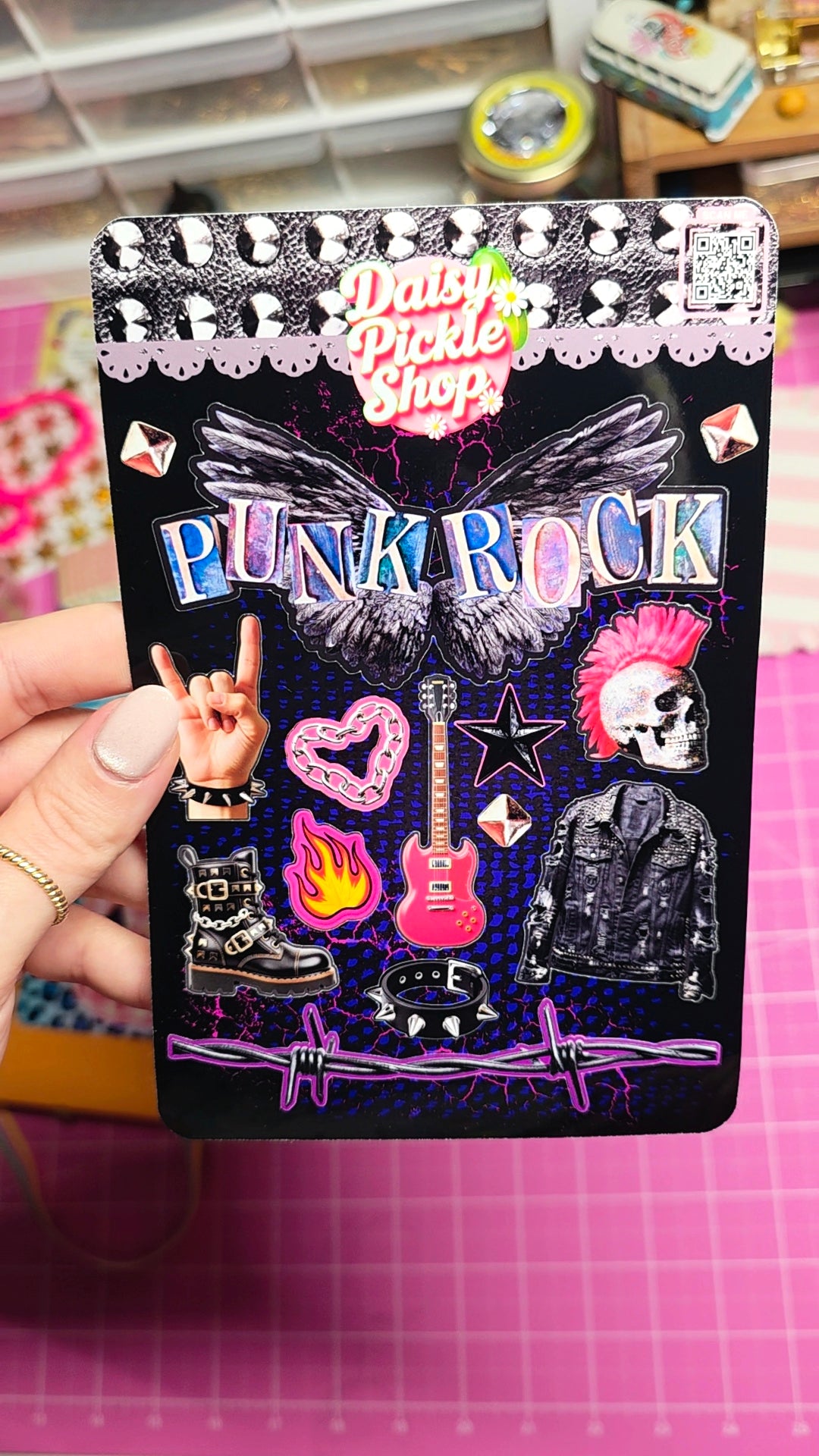 A hand holds a black enamel pin sheet featuring a punk rock theme with a wing, skull, boots, guitar, flame, and the text 'PUNK ROCK' and 'Daisy Pickle Shop'. The background shows a pink cutting mat and a craft room setting.