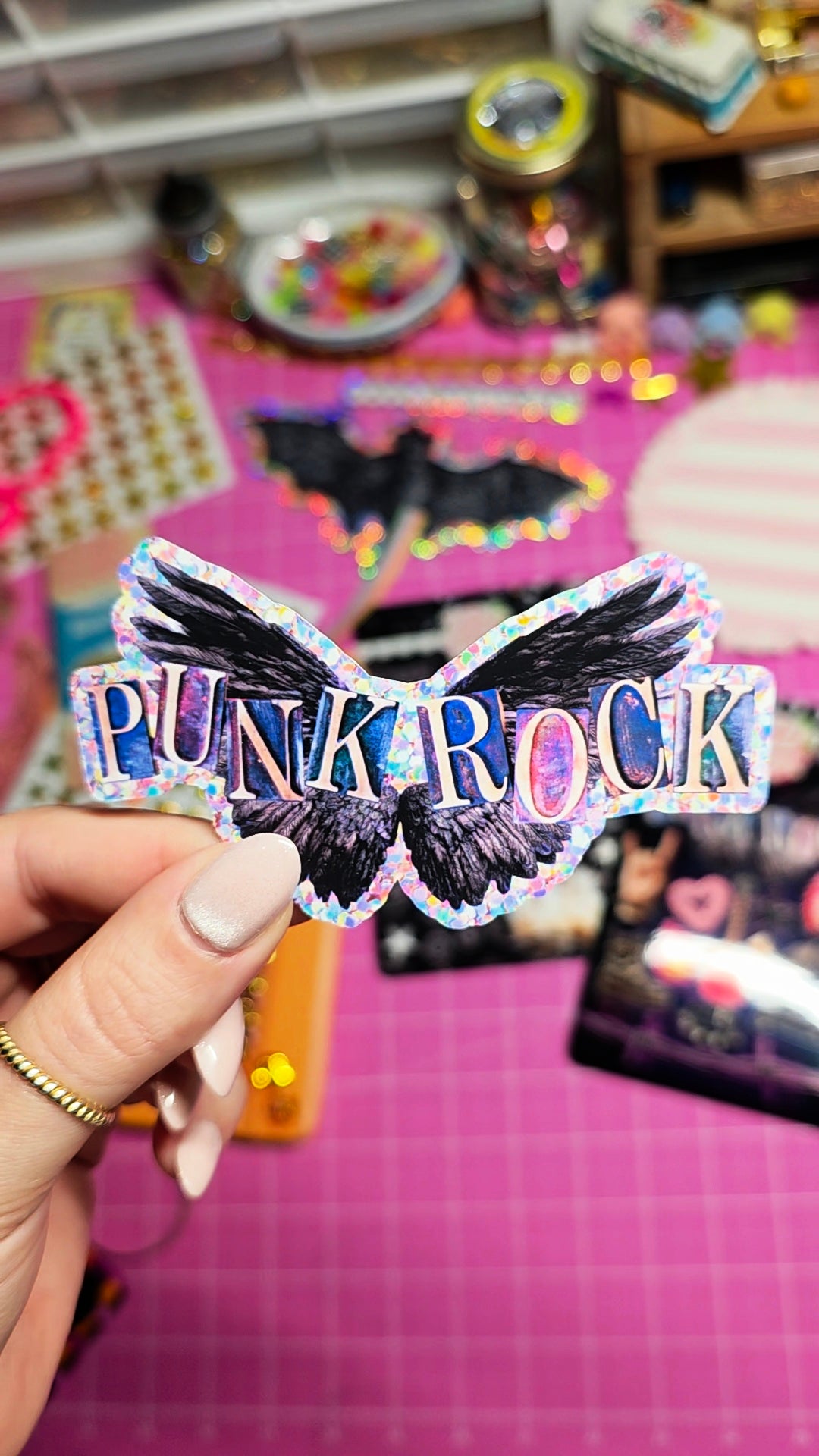 A hand holds a holographic sticker featuring a black raven with the text 'PUNK ROCK' in pink, purple, and blue glittery letters, set against a pink craft table background.