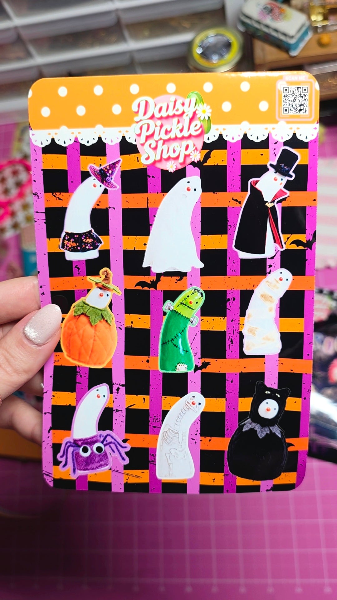 A hand holds a decorative tin sign featuring the 'Daisy Pickle Shop' logo and a Halloween-themed design with ghost, pumpkin, cat, spider, and mummy stickers on a black, pink, and orange striped background.