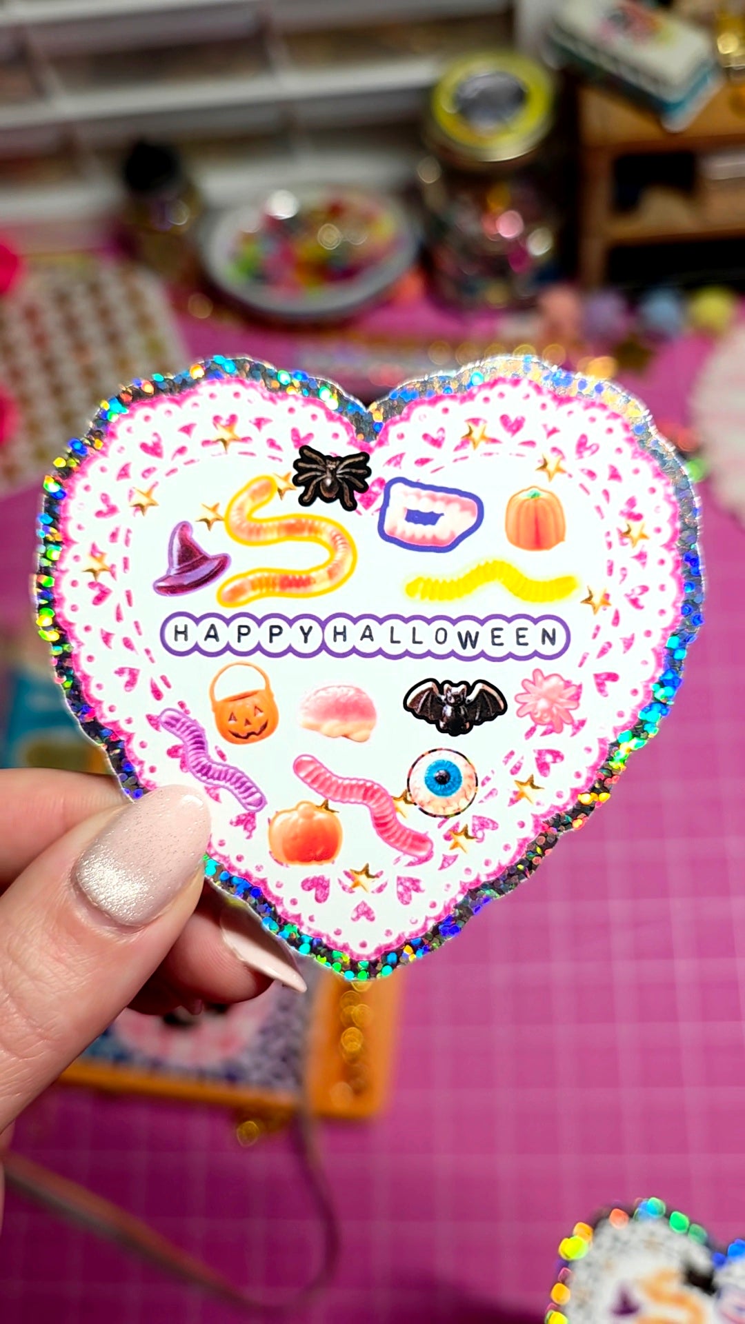 A hand holds a heart-shaped glittery card that says 'HAPPY HALLOWEEN' with cute Halloween illustrations like a witch hat, pumpkin, and spider on a pink background.
