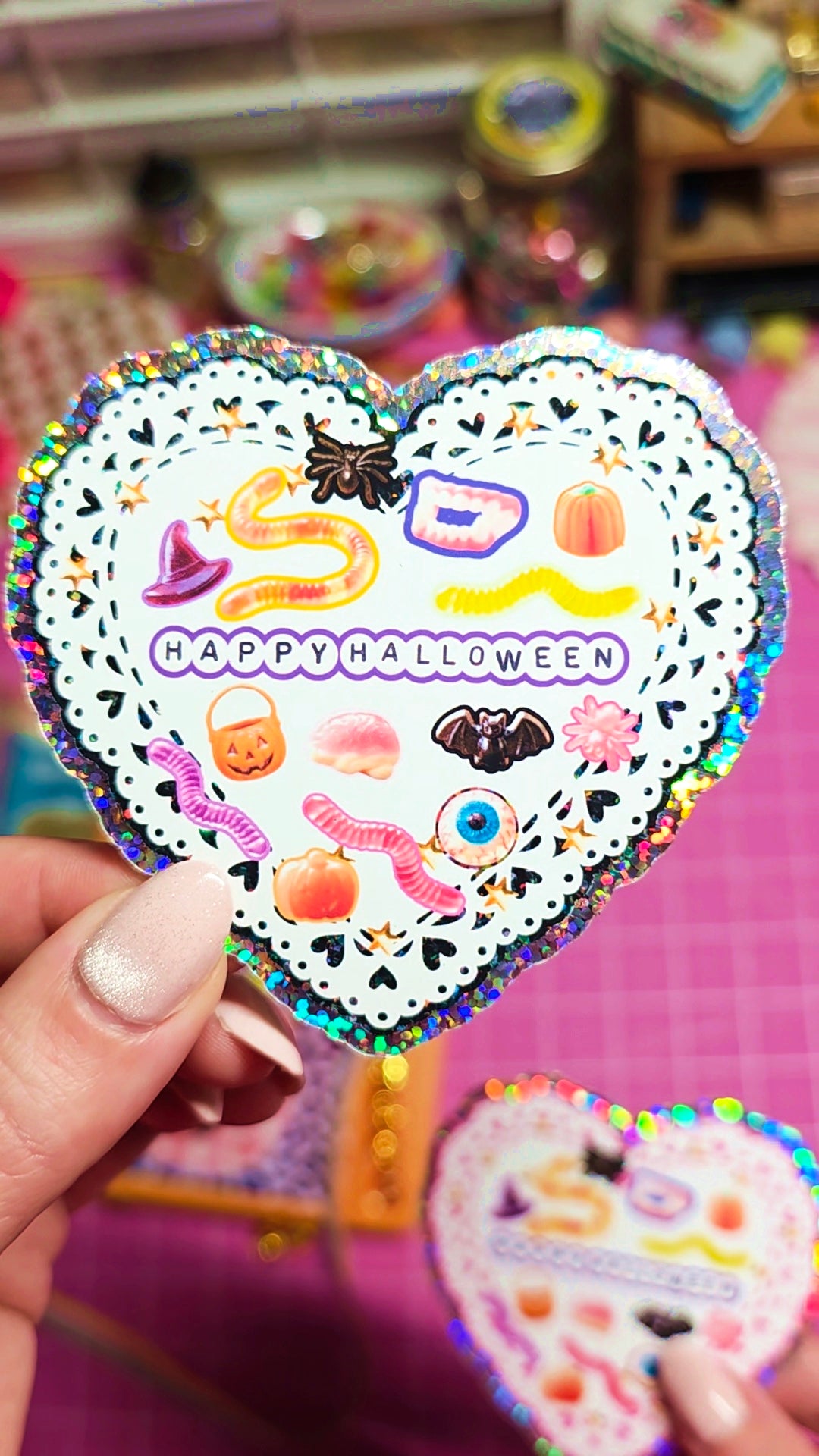 A hand holds a heart-shaped holographic sticker featuring a white lace pattern and colorful Halloween illustrations, including a witch hat, spider, pumpkin, and the words 'HAPPY HALLOWEEN'.