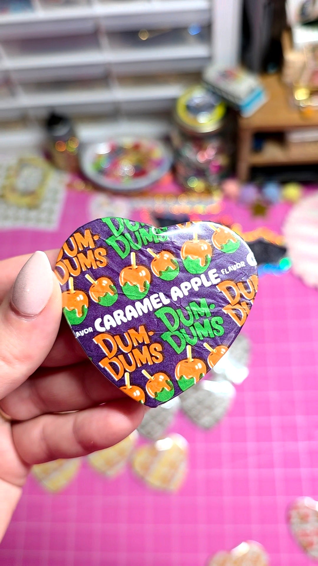 A hand holding a heart-shaped Dum Dums collectible pin featuring the Caramel Apple character, set against a pink craft mat with other pins and supplies blurred in the background.