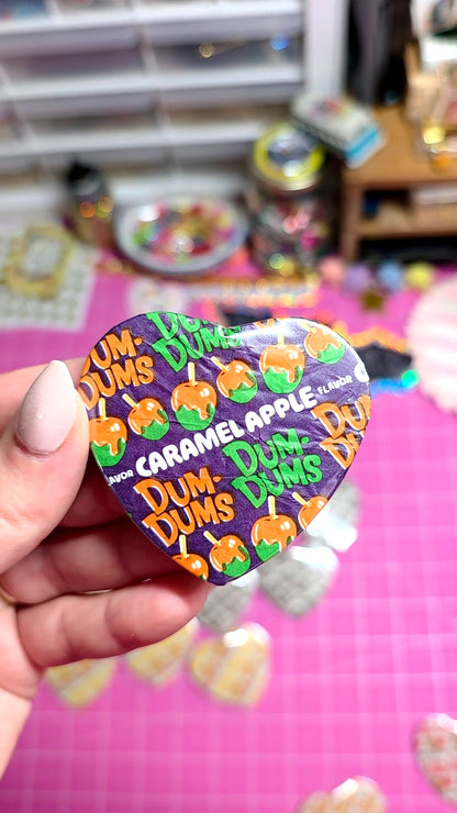 A hand holding a heart-shaped Dum Dums collectible pin featuring the Caramel Apple character, set against a pink craft mat with other pins and supplies blurred in the background.