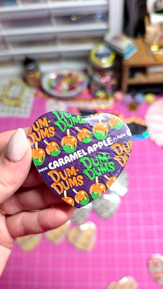 A hand holding a heart-shaped Dum Dums collectible pin featuring the Caramel Apple character, set against a pink craft mat with other pins and supplies blurred in the background.