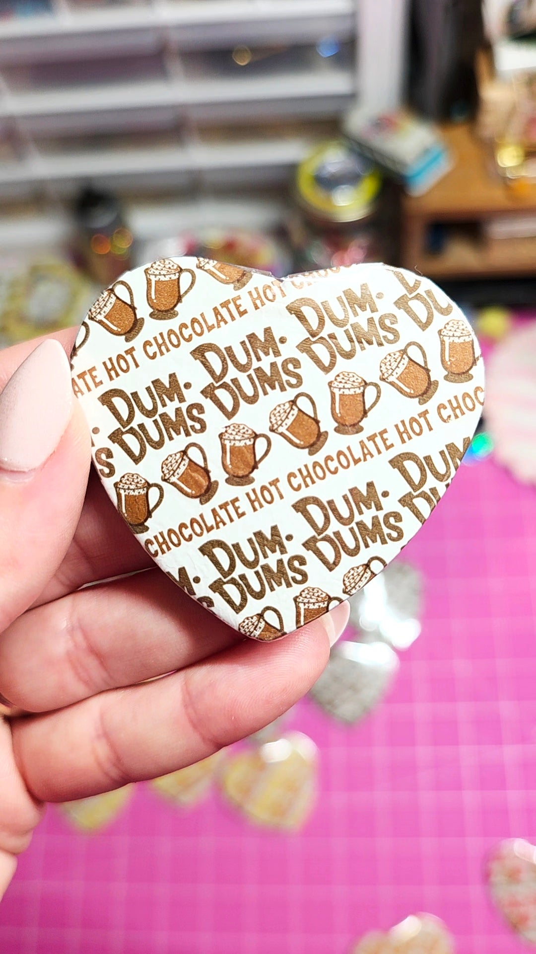 A hand holds a heart-shaped cookie or pin featuring a repeating pattern of brown hot chocolate mugs and the text 'DUM DUMS DUMS HOT CHOCOLATE'. The background is a pink cutting mat with other small heart-shaped items scattered around, and a blurry indoor setting in the distance.