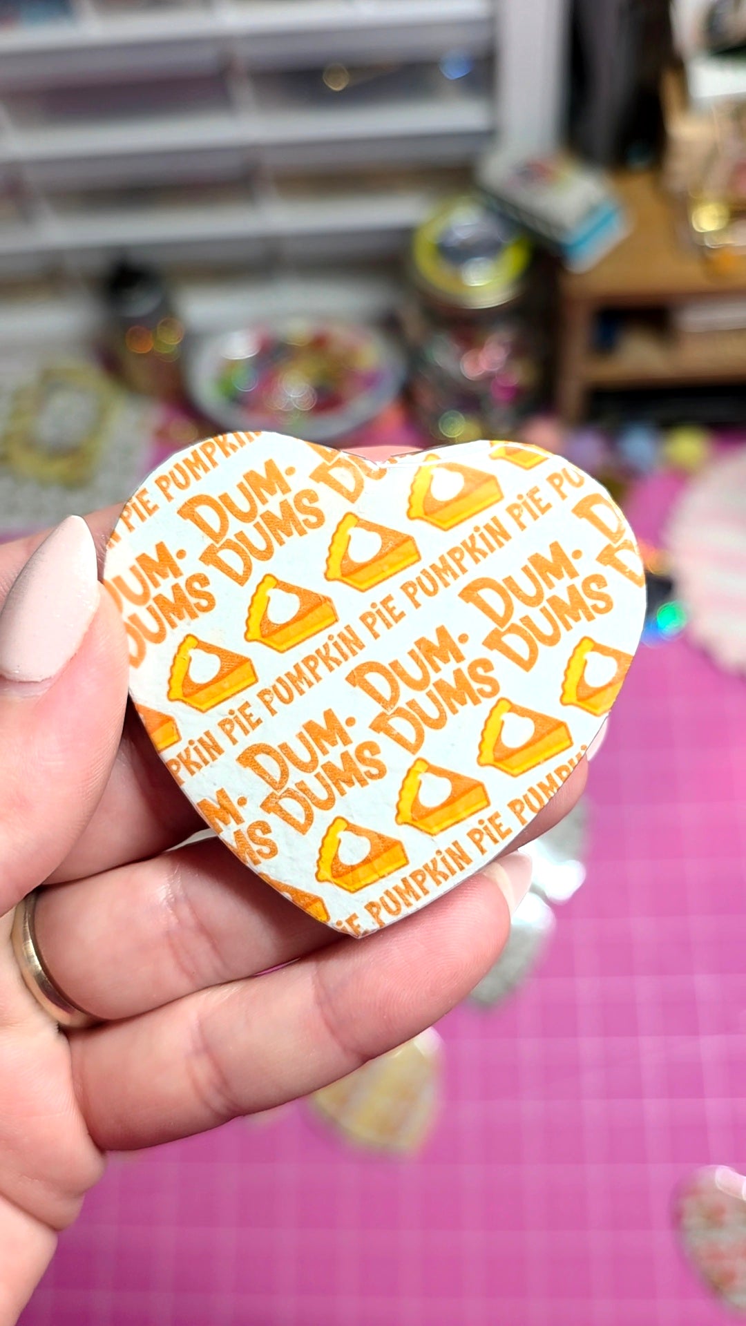 A hand holding a heart-shaped pin featuring a pumpkin pie design with the text 'Dum-Dum Pumpkin Pie' repeated in orange on a white background.