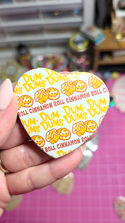A hand holding a heart-shaped pinback button featuring a white background with yellow 'Dum Dums' smiley faces and red 'Roll Cinnamon Roll' text, set against a pink cutting mat and a blurred craft room background.