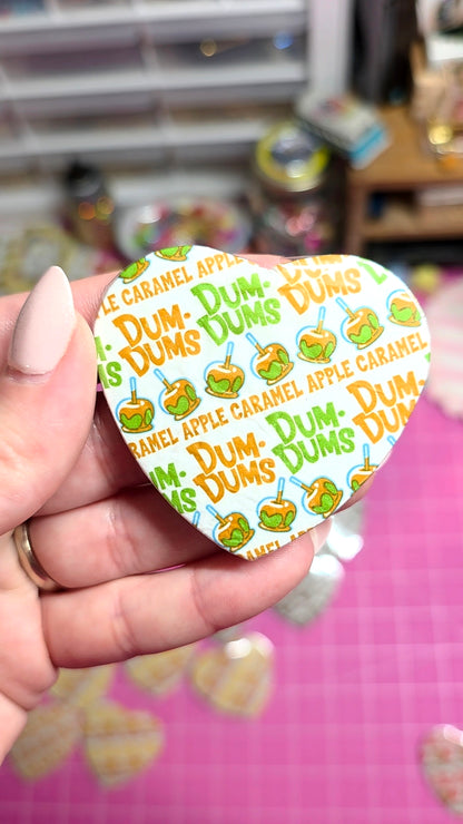 A hand with manicured nails holds a heart-shaped pin featuring a repeating 'Dum Dums' pattern with green and orange characters on a white background, with a pink cutting mat and blurred craft supplies in the background.