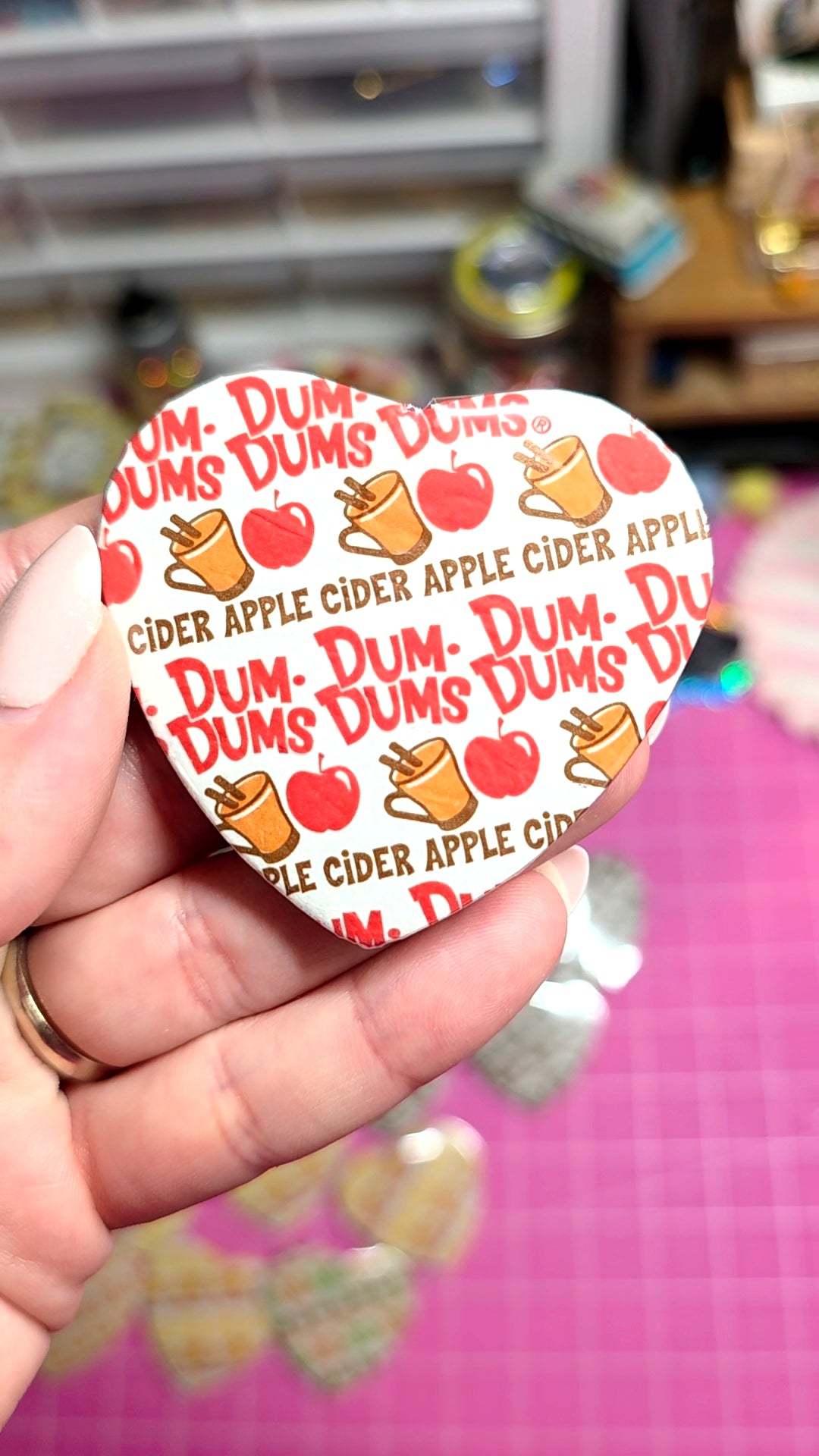 A hand holding a heart-shaped pinback button featuring a repeating pattern of red apples and orange coffee cups with the text 'CIDER APPLE CIDER'. The background is a pink cutting mat with other small pins visible in a blurred craft room setting.