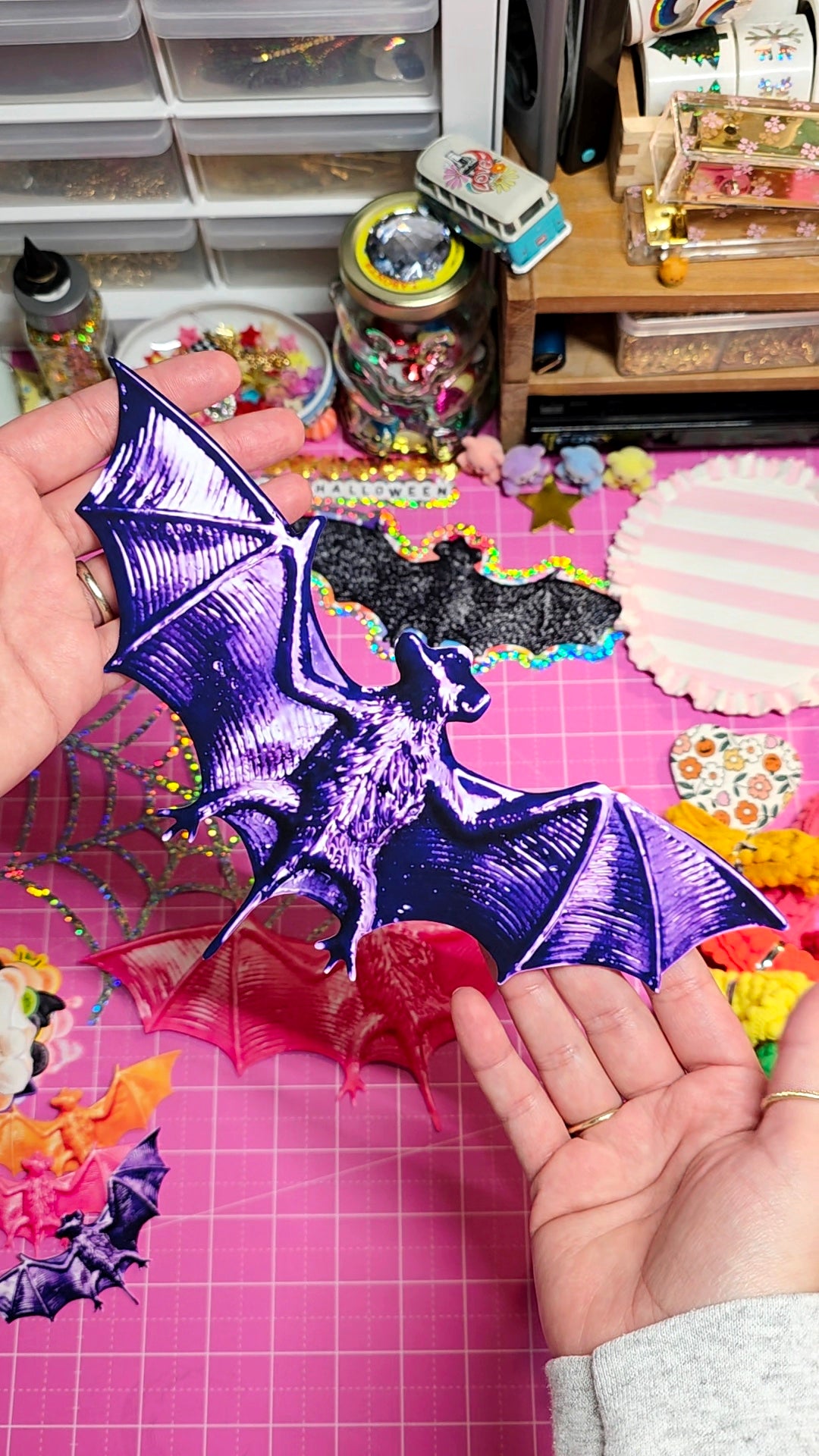 A person's hands holding a metallic purple bat-shaped paper cutout over a pink grid cutting mat in a craft room setting.