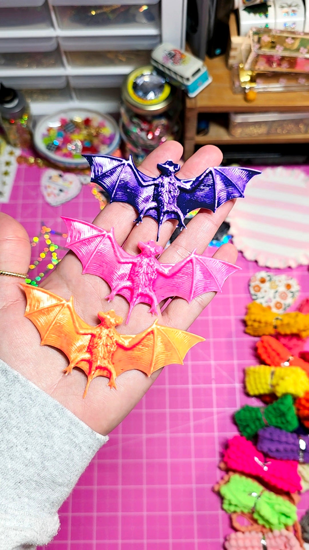 A hand holds three small bat-shaped accessories in purple, pink, and orange, with a pink craft mat and colorful accessories in the background.