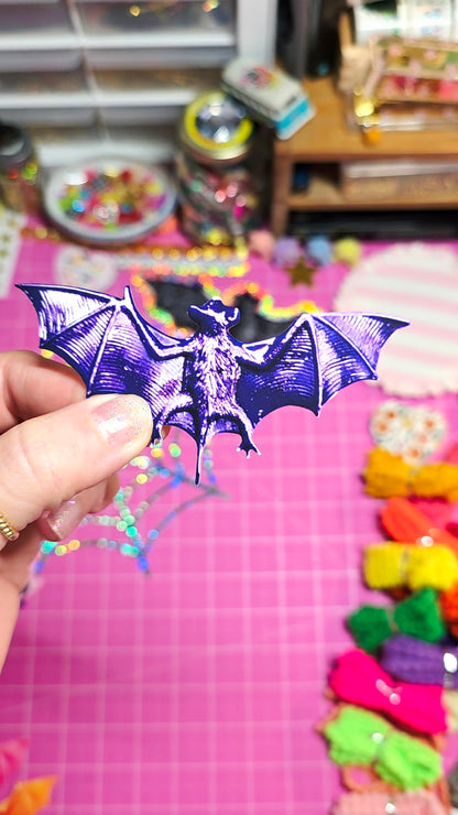 A hand holding a metallic purple bat-shaped accessory against a pink craft mat background with colorful accessories and blurred supplies.