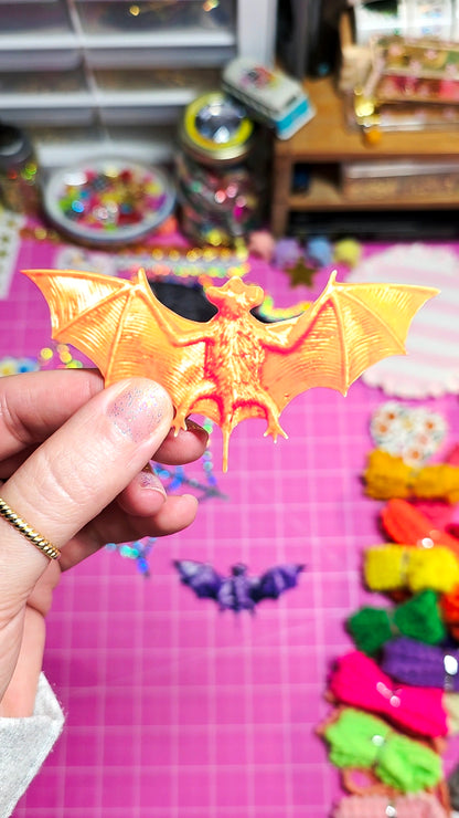 A hand holds a small orange bat-shaped accessory with spread wings against a pink craft mat background filled with colorful supplies.