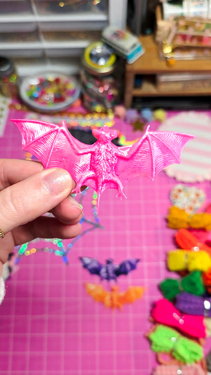 A hand holds a small pink bat-shaped accessory in front of a pink craft mat with colorful items in the background.