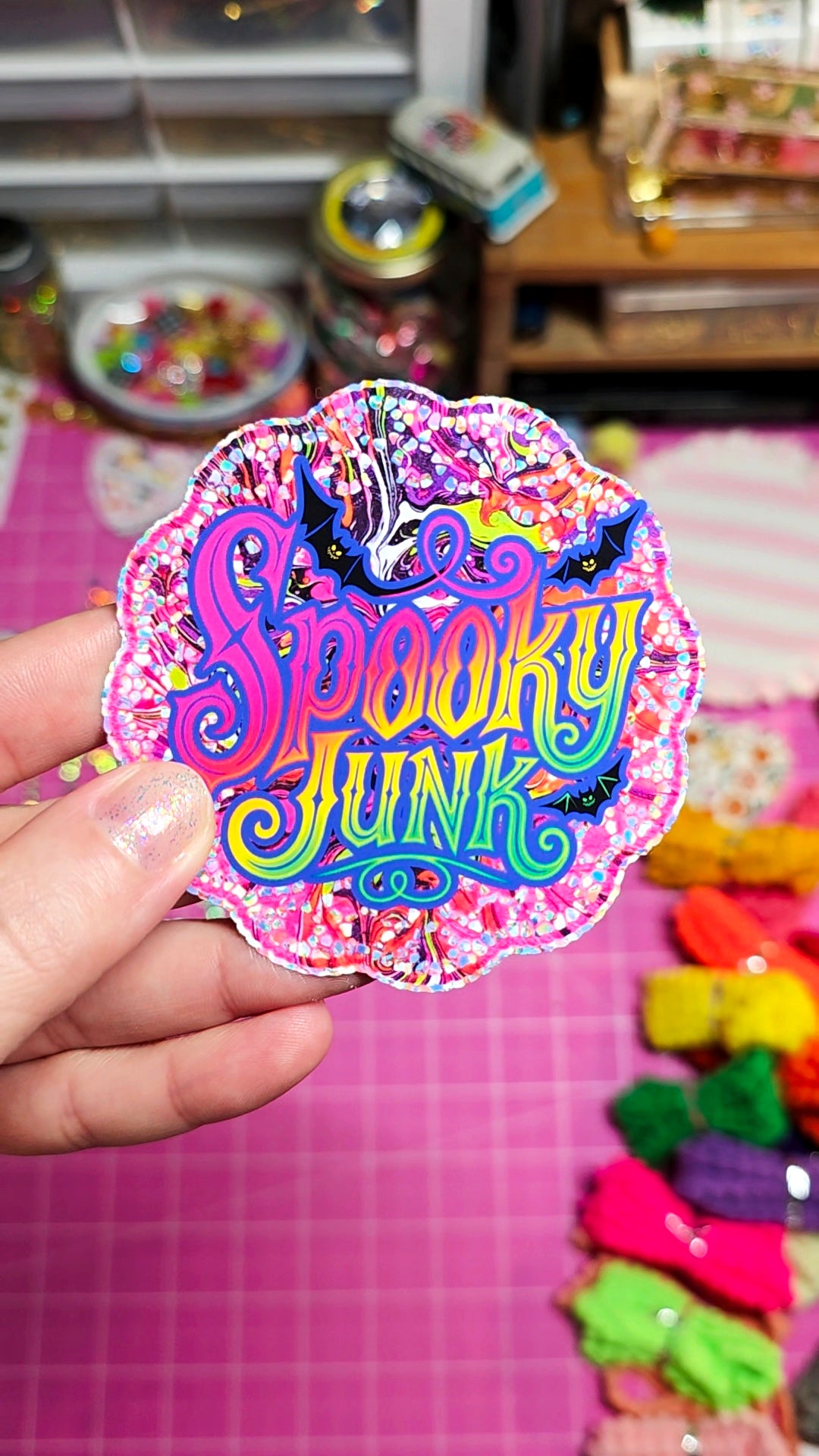A hand holds a scalloped, glittery sticker or patch featuring the words 'Spooky Junk' in colorful bubble letters with bats and a spider on a pink craft table background.