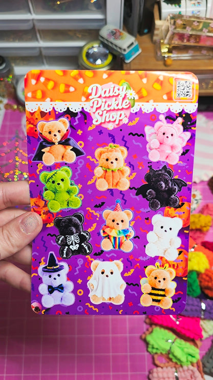 A hand holds a printed sticker sheet or card featuring cute Halloween-themed teddy bears and ghosts on a purple background with the text 'Daisy Pickle Shop.' The scene appears to be a craft or stationery workspace with colorful items blurred in the background.