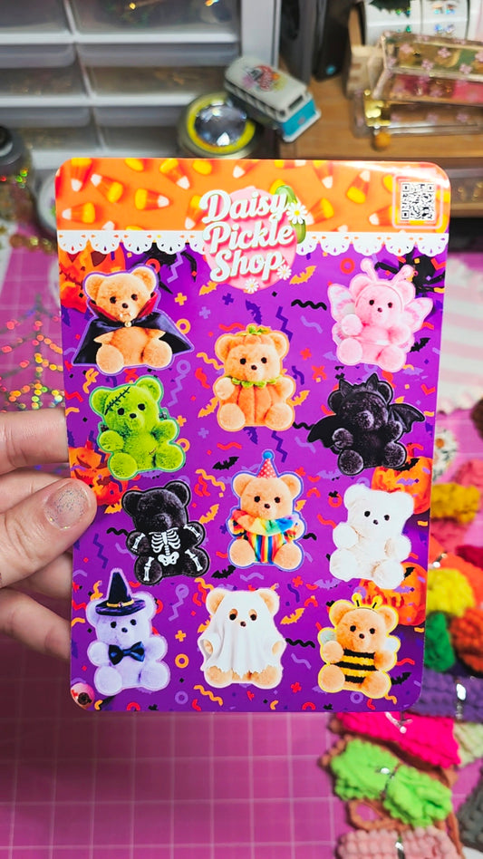 A hand holds a printed sticker sheet or card featuring cute Halloween-themed teddy bears and ghosts on a purple background with the text 'Daisy Pickle Shop.' The scene appears to be a craft or stationery workspace with colorful items blurred in the background.