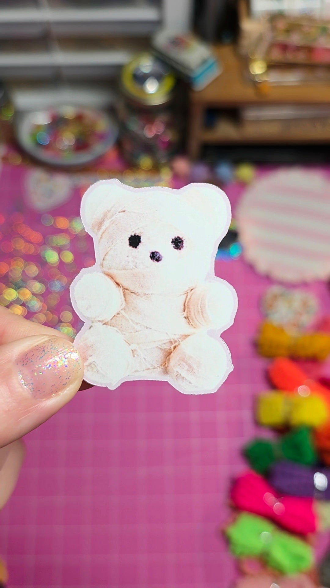 A small plush teddy bear accessory held by a hand over a pink craft mat with colorful supplies in the background.