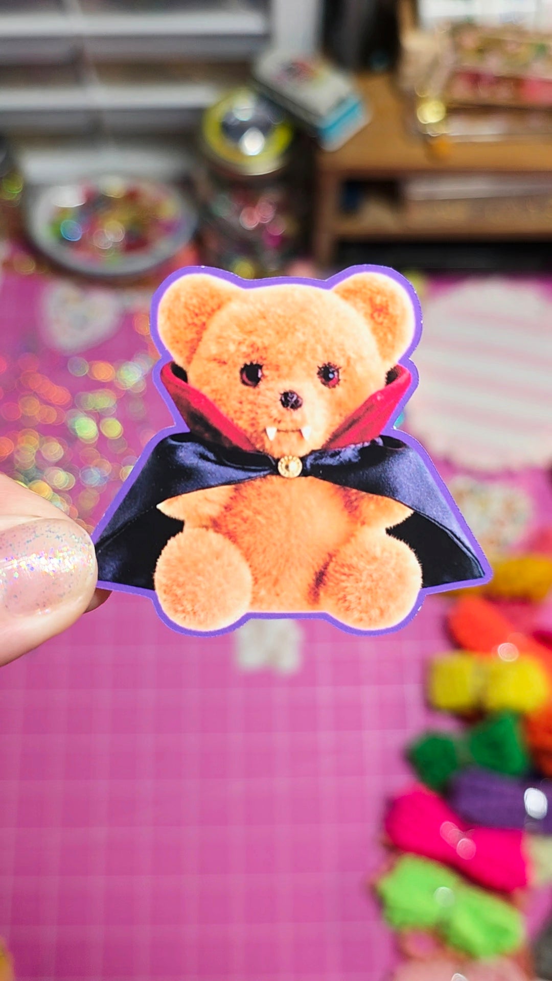 A hand holds a die-cut sticker or patch featuring a fluffy orange teddy bear wearing a black cape with a red collar and a small gold bell, set against a pink craft workspace background.