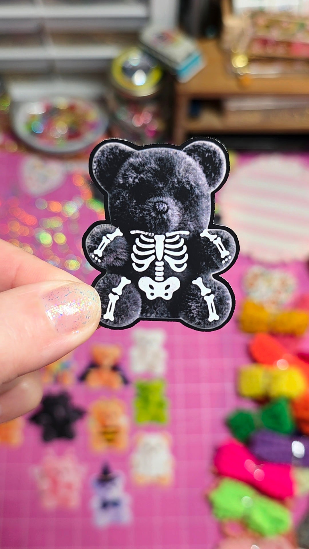 A hand holds a black gummy bear sticker or patch featuring a white skeleton print, with a colorful blurred background of small craft items and gummy bears on a pink surface.