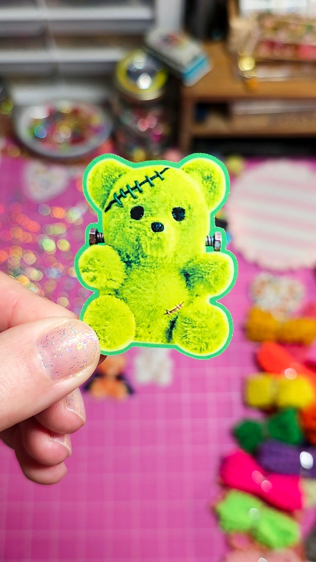 A hand holding a bright lime green teddy bear hair clip with black stitched details and small black accents, set against a colorful pink background with other clips.