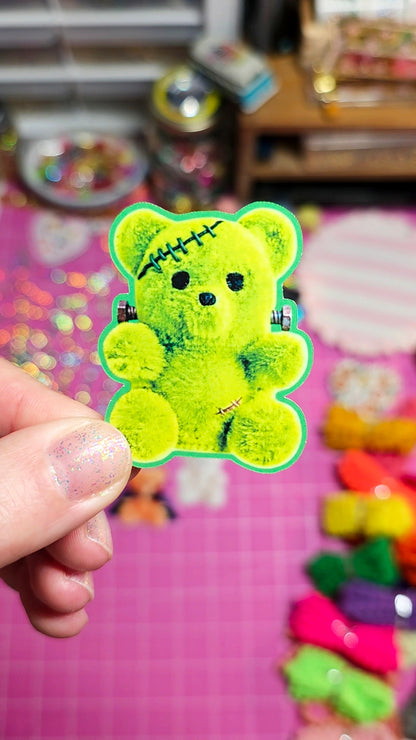 A hand holding a bright lime green teddy bear hair clip with black stitched details and small black accents, set against a colorful pink background with other clips.