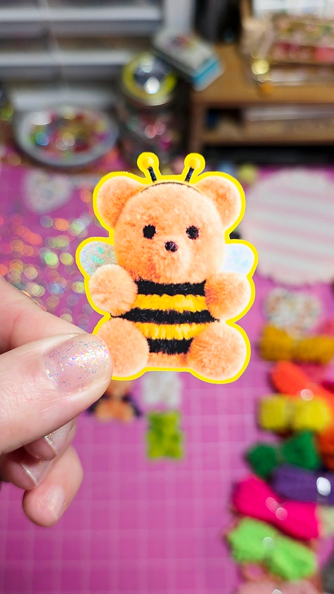A hand holds a small embroidered bee-shaped patch or applique with a fuzzy orange teddy bear body, yellow and black striped body, and yellow wings, against a colorful blurred craft background.