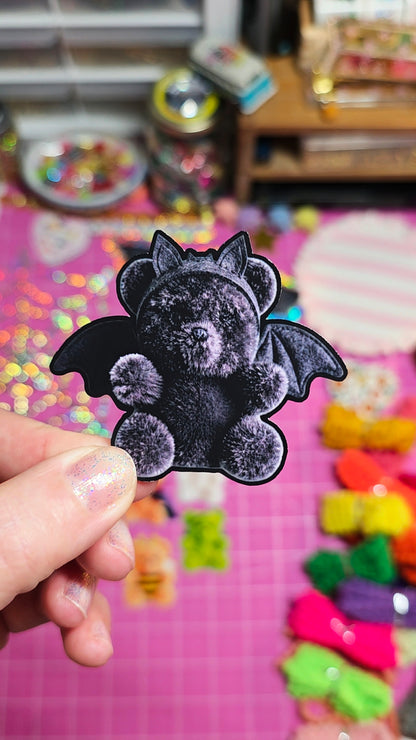 A hand holds a black bat-shaped teddy bear patch or sticker with outstretched wings against a pink craft table filled with colorful embellishments and small items.